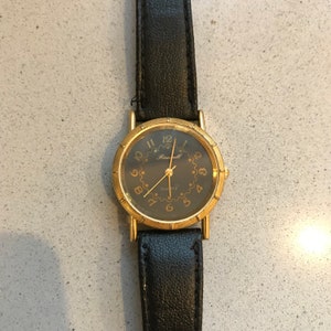 Op de afbeelding: Een gouden polshorloge met een zwarte leren band en een zwarte wijzerplaat. De wijzerplaat van het horloge heeft gouden uurmarkeringen en wijzers, en het woord "Rockwell" is in goud bovenaan de wijzerplaat gedrukt. Het horloge heeft ook een klein gouden "Quartz"-label onderaan de wijzerplaat.