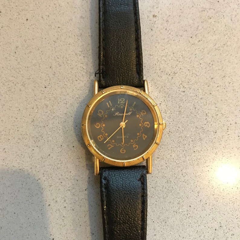 Op de afbeelding: Een gouden polshorloge met een zwarte leren band en een zwarte wijzerplaat. De wijzerplaat van het horloge heeft gouden uurmarkeringen en wijzers, en het woord "Rockwell" is in goud bovenaan de wijzerplaat gedrukt. Het horloge heeft ook een klein gouden "Quartz"-label onderaan de wijzerplaat.