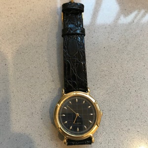 Op de afbeelding: Een gouden polshorloge met een zwarte wijzerplaat en een zwarte leren band. De wijzerplaat van het horloge heeft een rasterpatroon en het woord "Quartz" staat erop gedrukt.