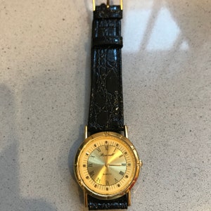 Op de afbeelding: Een gouden polshorloge met Romeinse cijfers voor de uren en een zwarte leren band. De wijzerplaat van het horloge zegt "Roswell Quartz".