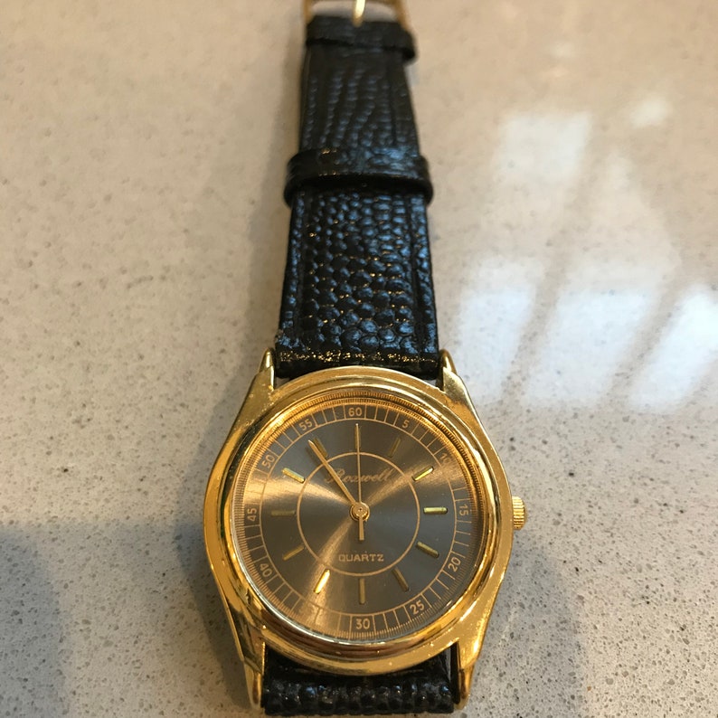 Op de afbeelding: Een gouden polshorloge met een zwarte wijzerplaat en een zwarte leren band. De wijzerplaat van het horloge heeft gouden uurmarkeringen en wijzers, en het woord "Rowell" staat in het midden gedrukt. De wijzerplaat van het horloge heeft ook het woord "QUARTZ" gedrukt onder de merknaam.