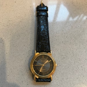Op de afbeelding: Een gouden polshorloge met een zwarte leren band en een zwarte wijzerplaat. De wijzerplaat van het horloge heeft een gouden bezel en gouden Romeinse cijfers als uurmarkeringen. De wijzerplaat van het horloge heeft ook een gouden secondewijzer en een gouden minutenwijzer. Op de wijzerplaat van het horloge staat "Rosendahl". Op de wijzerplaat van het horloge staat ook "QUARTZ".