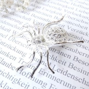 Peut inclure: Broche araignée argentée avec un corps orné de cristaux étincelants et des pattes délicates. La broche est posée sur un fond de texte blanc et ornée de petites fleurs blanches, créant un accessoire fantaisiste et élégant.