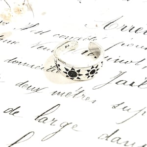 Peut inclure: Une bague en argent avec un motif de soleil noir. La bague est posée sur une surface blanche avec du texte manuscrit.