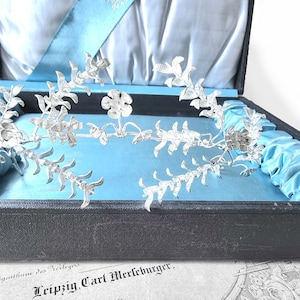 May include: A silver floral design napkin ring in a blue velvet lined box. The box has the text "Eigentum des Verlegers Leipzig, Carl Merkleburger." on the bottom.