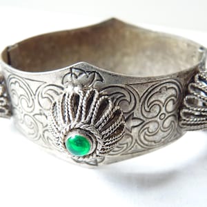 Può includere: Un braccialetto d'argento con un designo intricato e tre pietre preziose verdi. Il braccialetto è fatto di metallo e ha un motivo decorativo.