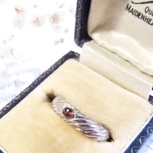 Può includere: Un anello d'argento con una pietra preziosa rossa incastonata al centro. L'anello ha una fascia testurizzata ed è esposto in una scatola per anelli in velluto nero.