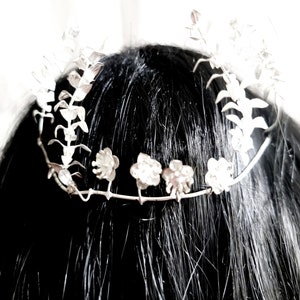 May include: A silver floral crown with delicate white flowers and leaves. The crown is made of wire and is designed to be worn on the head.