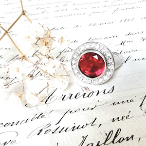 Peut inclure: Gros plan d'une bague en argent avec une grande pierre précieuse rouge entourée de petites pierres précieuses blanches. La bague est posée sur un morceau de papier avec une écriture cursive.