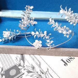 May include: A silver floral headband with delicate leaves and flowers. The headband is displayed in a blue velvet jewelry box.