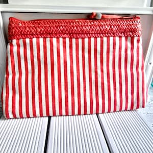 Op de afbeelding: Een rood-wit gestreepte clutch met een geweven rode bovenkant. De tas heeft een ritssluiting en een kleine lus. De tas is rechthoekig en lijkt van stof te zijn.