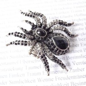 Peut inclure: Broche araignée détaillée avec un corps et des pattes en métal argenté, ornée de cristaux noirs et transparents. L'araignée a deux grands yeux en pierres noires et une gemme en forme de goutte sur l'abdomen.