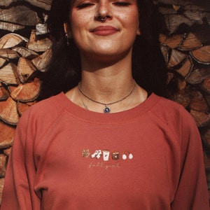 Könnte beinhalten: Rostfarbenes Sweatshirt mit gestickten Herbstmotiven und dem Text "fall girl". Das Design umfasst ein Blatt, Noten, eine Kaffeetasse, eine Kerze und Kopfhörer. Das Sweatshirt hat einen Rundhalsausschnitt.