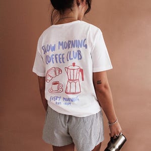 Könnte beinhalten: Weißes T-Shirt mit einem blauen und roten Grafikdesign, auf dem "Slow Morning Coffee Club" steht und ein Croissant, eine Kaffeekanne und eine Kaffeetasse abgebildet sind. Der Text unter der Grafik lautet "Every Morning 7am-10am".