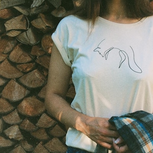 Könnte beinhalten: Ein weißes T-Shirt mit einer einfachen Linienzeichnung eines Fuchses auf der Vorderseite. Das Shirt wird von einer Person getragen, die vor einem Stapel Brennholz steht.