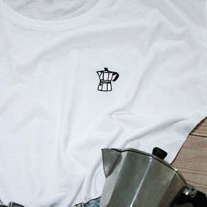 Könnte beinhalten: Weißes T-Shirt mit einem kleinen, schwarz gestickten Kaffeekannen-Design. Das Shirt besteht aus einem weichen Material und wird mit Jeans kombiniert. Eine silberne Kaffeemaschine ist ebenfalls im Bild.