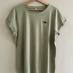 Könnte beinhalten: Ein hellgrünes T-Shirt mit einem kleinen gestickten Bild eines Otters auf der Vorderseite.