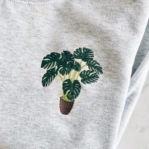Könnte beinhalten: Ein graues Sweatshirt mit einer gestickten grünen Monstera-Pflanze in einem braunen Topf.