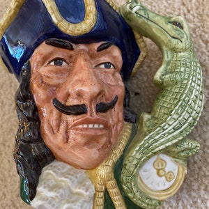 May include: A ceramic mug shaped like a pirate's head with a blue hat and a green alligator handle. The pirate has a mustache and a beard.