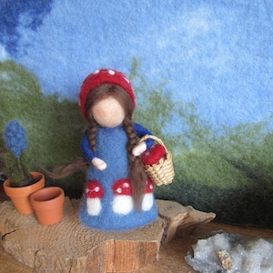 Könnte beinhalten: Eine Filzpuppe in einem blauen Kleid mit weißen und roten Pilzverzierungen, einem roten Pilzhut und Zöpfen. Die Puppe hält einen kleinen Korb mit roten Gegenständen. Der Hintergrund zeigt eine blaue und grüne Filzlandschaft.