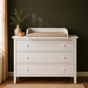 Op de afbeelding: Een witte commode met drie lades en een aankleedtafelopzetstuk. De aankleedtafel heeft een beige kussen. De commode heeft drie lades met ronde knoppen. Een vaas met groen en een klein geweven mandje staan bovenop.