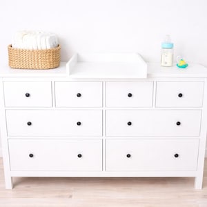 Op de afbeelding: Een witte commode met zwarte knoppen, een aankleedkussen, een rieten mand met luiers, een babyfles en een speen. De commode heeft meerdere lades en een platte bovenkant, perfect voor een babykamer.