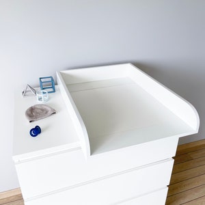 Op de afbeelding: Witte commode met vier lades en een witte verschoningsmat. De verschoningsmat heeft een verhoogde rand en is lichtjes gekanteld. Er liggen een paar kleine voorwerpen op de commode, waaronder een muts, een speen en een paar kleine speelgoedjes.