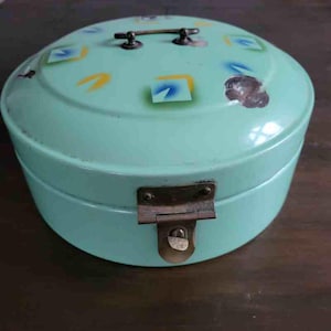 Op de afbeelding: Een vintage, ronde, mintgroene metalen doos met een scharnierend deksel en een sluiting. Het deksel heeft een handvat en is versierd met geometrische vormen in geel, blauw en wit. De doos staat op een donker houten oppervlak.