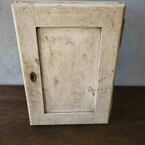 Op de afbeelding: Een kleine, antieke houten kast met een enkele deur. De kast is geschilderd in een verweerde gebroken witte kleur, met tekenen van slijtage en ouderdom. Een klein, rond sleutelgat is zichtbaar op de deur. De kast wordt waarschijnlijk gebruikt voor opslag.