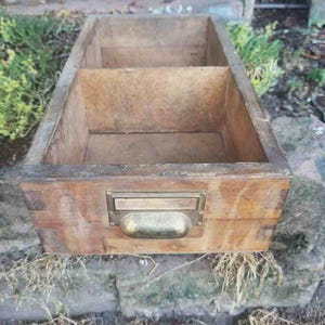 Peut inclure: Un tiroir rectangulaire en bois avec une finition brune patinée et des assemblages à queue d'aronde visibles. La façade comprend une poignée cuvette en métal doré et un porte-étiquette rectangulaire. L'intérieur est divisé en deux sections.