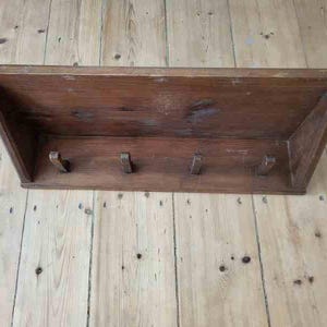 Op de afbeelding: Een rechthoekige houten wandplank met een plat bovenoppervlak en een verticaal achterpaneel. De unit bevat vier kleine, gelijkmatig verdeelde houten haken bevestigd aan het onderste horizontale gedeelte, alles afgewerkt in een middelbruine houttint.