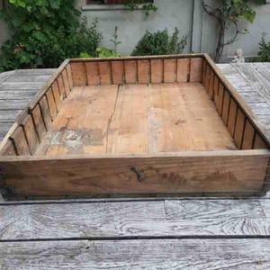 Può includere: Una cassa di legno rettangolare con la parte superiore aperta, dall'aspetto invecchiato. La cassa è costruita in legno chiaro, con venature visibili e una patina leggermente invecchiata. I lati sono rinforzati con doghe verticali e si vede un fermo metallico.