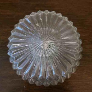 Könnte beinhalten: Eine klare Glasschale mit einem strukturierten, geometrischen Design. Die Schale hat eine kreisförmige Form mit einer Reihe von abgewinkelten Rippen, die von der Mitte ausgehen. Die Schale steht auf einer dunkelbraunen Holzoberfläche.
