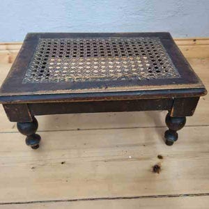 Può includere: Uno sgabello basso rettangolare con una struttura in legno marrone scuro e un pannello centrale intarsiato con un motivo a intreccio di canna. Lo sgabello poggia su quattro gambe corte, arrotondate e tornite, con segni visibili di usura.