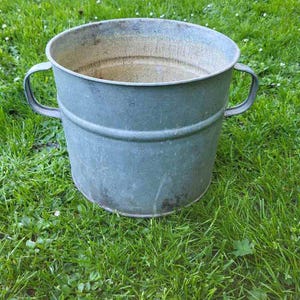 Op de afbeelding: Een verweerde, blauwgroene metalen emmer met twee gebogen handvatten. De emmer heeft een cilindrische vorm met een licht uitlopende rand. Hij staat op een bed van groen gras.