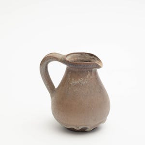 Könnte beinhalten: Ein kleiner, brauner Keramik Krug mit einem gesprenkelten Überzug. Der Krug hat einen runden Körper und einen gebogenen Griff.