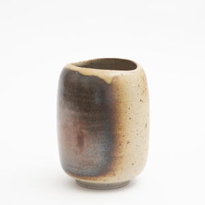 Può includere: Un piccolo vaso cilindrico in ceramica con una smaltatura marrone, beige e grigia. Il vaso ha un fondo leggermente arrotondato e un'ampia apertura.