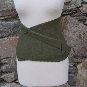Op de afbeelding: Een groene stoffen riem met een kruislings ontwerp. De riem wordt getoond op een wit mannequins tegen een stenen achtergrond.
