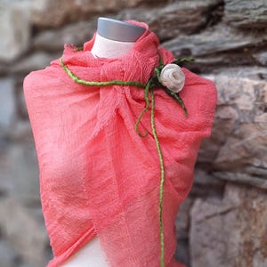 Puede incluir: Bufanda color coral drapeada sobre un maniquí, adornada con una rosa de tela blanca y detalles verdes. La bufanda tiene una apariencia texturizada y se encuentra sobre un fondo de pared de piedra.