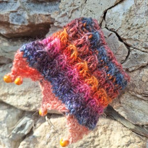 Op de afbeelding: Een handgebreid item met een textuurpatroon. Het heeft een levendige mix van oranje, roze, paarse en blauwe garens. Kleine, oranje kralen zijn aan de randen bevestigd. Het item wordt gepresenteerd tegen een stenen achtergrond.