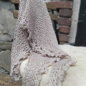 Op de afbeelding: Een delicate, lichtbeige gebreide sjaal met een kantrand, gedrapeerd over een stoel. De sjaal is gemaakt van een zacht, lichtgewicht garen en heeft een vloeiende, etherische uitstraling.