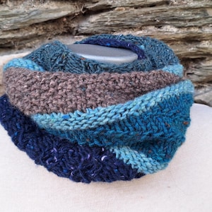 Puede incluir: Un cuello tejido a mano en tonos de azul, verde azulado y marrón, exhibido en un maniquí blanco. La prenda de punto texturizada presenta rayas de diferentes anchos y tonos, creando un accesorio acogedor y elegante. El fondo es una pared de piedra.