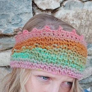 Op de afbeelding: Een veelkleurig gehaakt hoofdband met een kroonvormige bovenkant. De hoofdband is gemaakt van roze, oranje, groen en bruin garen.