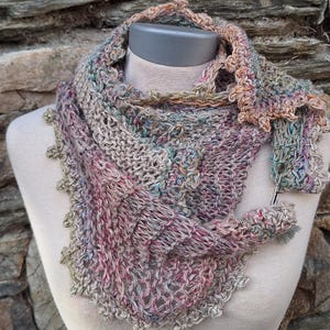 Puede incluir: Bufanda tejida a mano, multicolor, exhibida en un maniquí. Presenta una mezcla de hilos rosas, grises y azules, con un delicado borde festoneado. Una pequeña pieza tejida a juego está sujeta a la bufanda con un alfiler plateado.