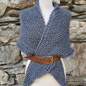 Op de afbeelding: Een grijze gebreide sjaal met een bruine leren riem. De sjaal is gedrapeerd over een wit mannequin en wordt getoond tegen een stenen muur.