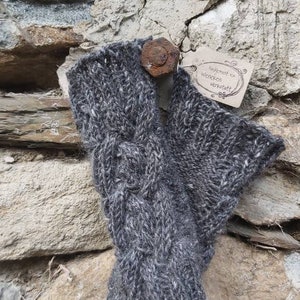 May include: Grey knitted fingerless gloves with a cable knit pattern. The gloves are made from wool and have a rustic look. The tag attached to the gloves reads "Handgemacht von Wockenwerkstatt".