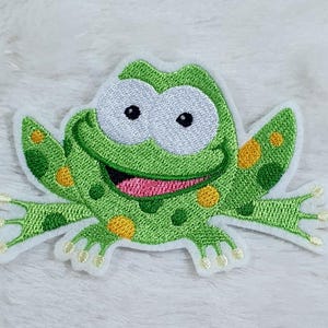 Könnte beinhalten: Gestickter Aufnäher mit einer Cartoon-Frosch-Figur mit großen weißen Augen, einem rosa Mund und grüner Haut mit gelben Punkten. Der Frosch hat ausgestreckte Arme und Beine und ist weiß umrandet. Geeignet für Kleidung oder Accessoires.