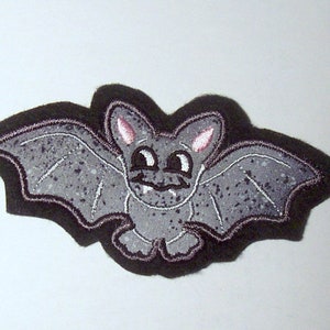 Könnte beinhalten: Eine graue Filzfledermaus mit rosa Ohren und weißen Augen. Die Fledermaus hat ein gesprenkeltes graues Muster auf ihren Flügeln.