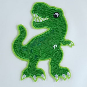 Könnte beinhalten: Grün gestickter Bügelaufkleber mit einem Cartoon-Dinosaurier mit weißen Zähnen und einem schwarzen Auge.