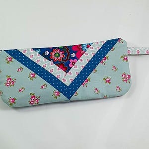 Könnte beinhalten: Eine hellblaue Reißverschlusstasche mit einem Blumenmuster und einem blau-weißen geometrischen Design. Die Tasche hat einen kleinen blauen Reißverschluss-Zipper und einen blau-weißen gemusterten Riemen.